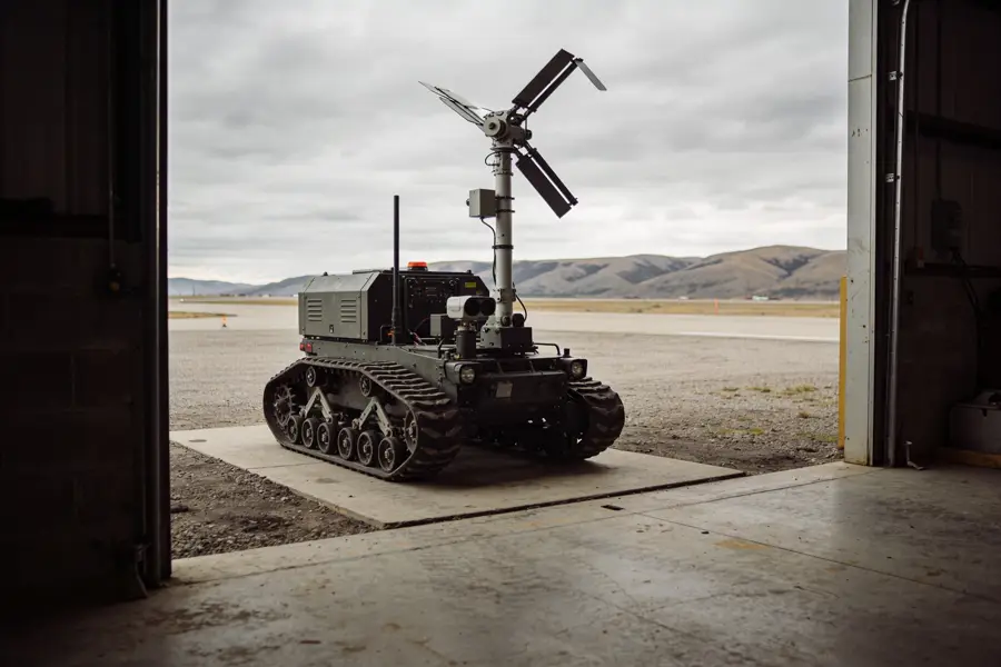 A tracked uncrewed ground vehicle parked inside an open hangar door, foothills beyond.