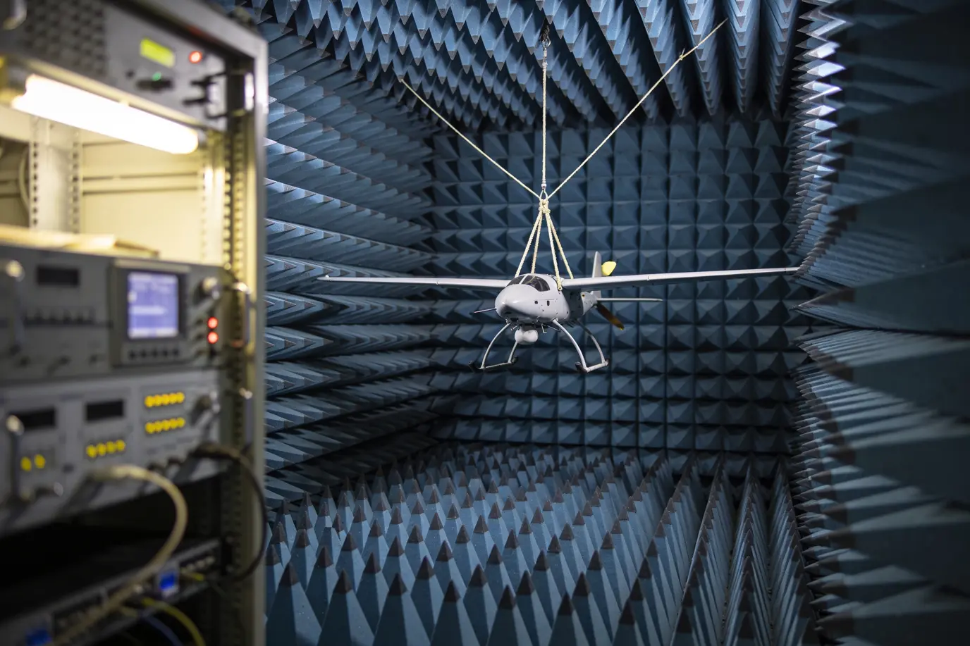 A small uncrewed aircraft suspended on a rig at the centre of an anechoic test chamber lined with blue-grey absorber foam.